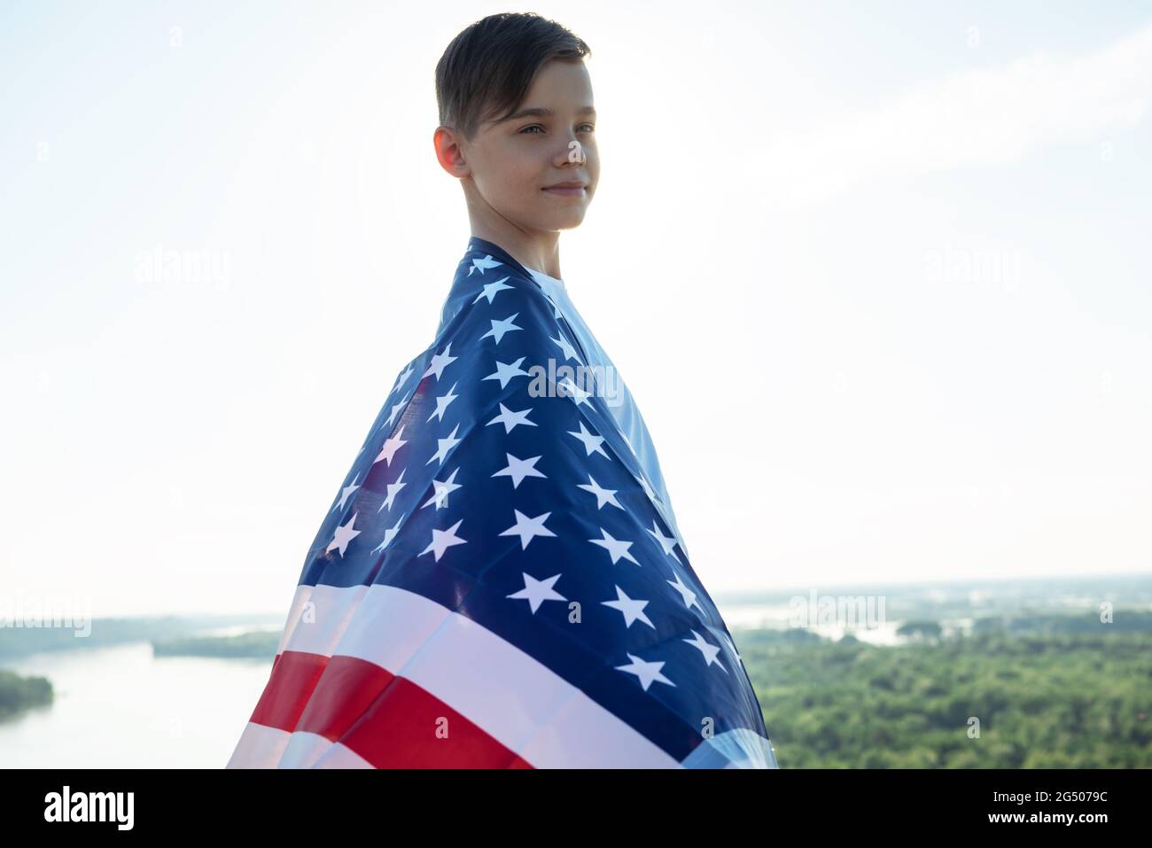 Blonde boy waving national USA flag outdoors over blue sky at the river bank. Beauty summer sunny day. American flag, patriotism, independence day 4th Stock Photo