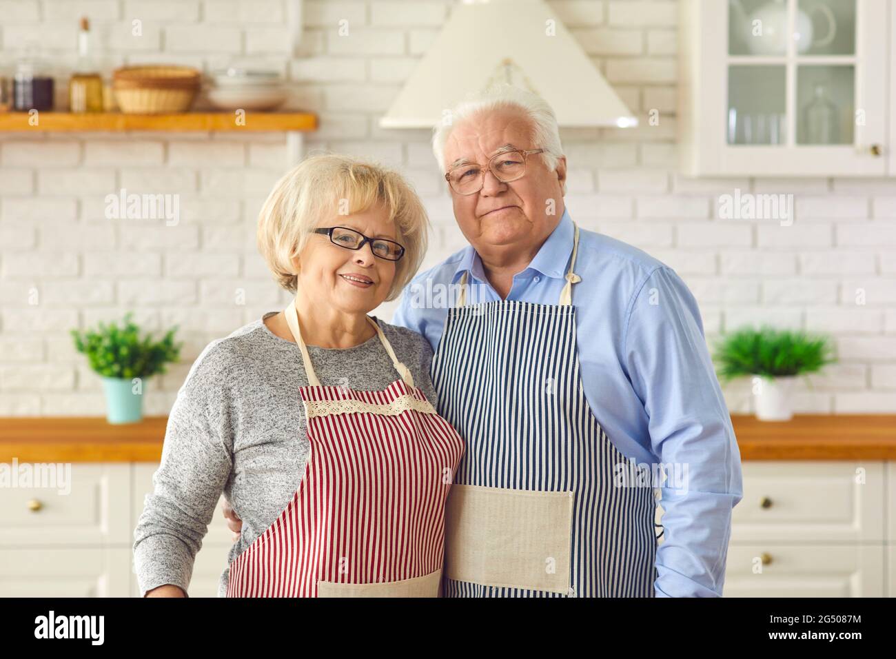 Portrait of a happy married senior couple dressed in aprons and posing in their kitchen. Stock Photo