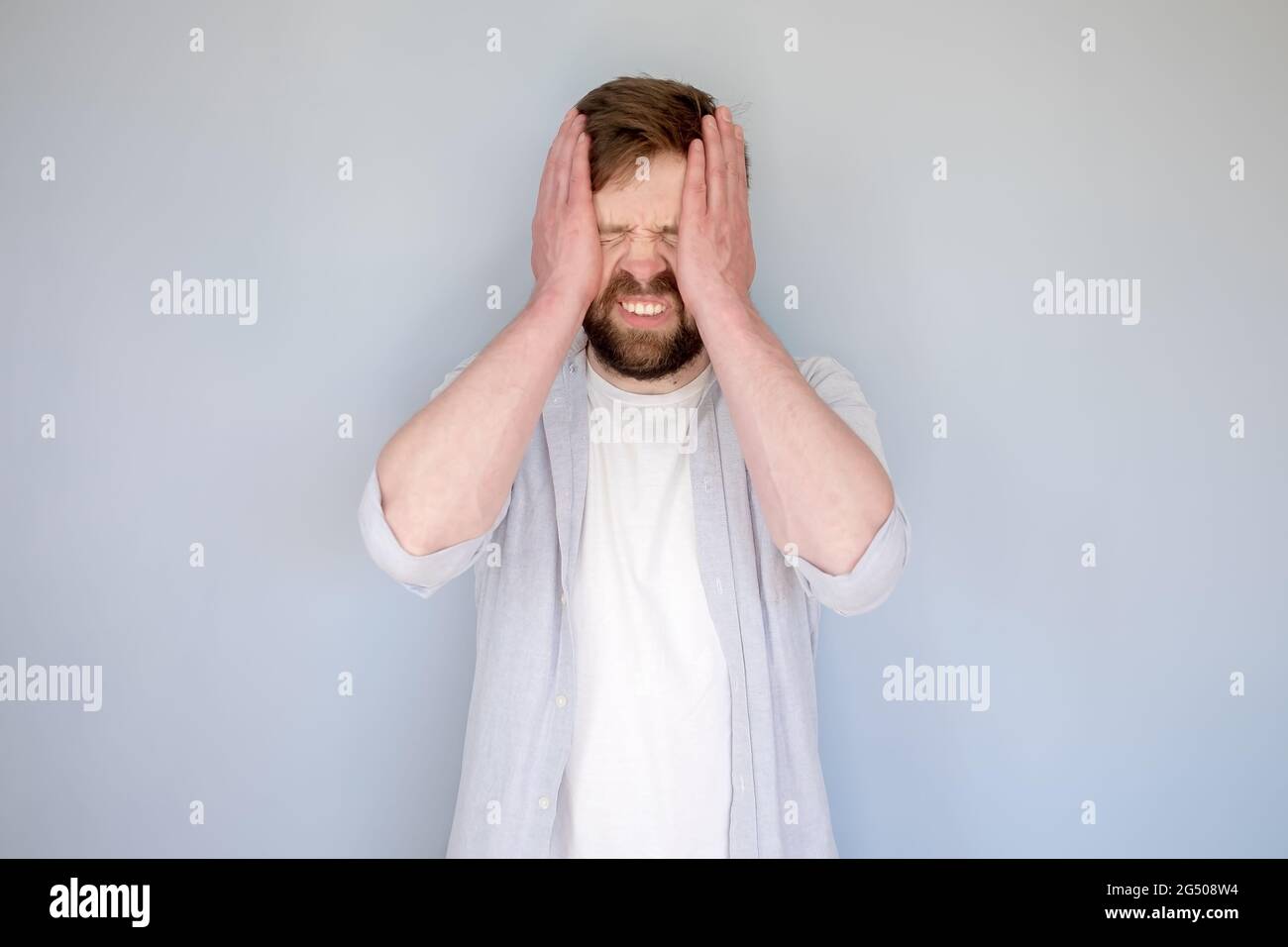 Caucasian man in stress or suffering from headache, he closed his eyes, holds head with hands and bares teeth. Gray background. Stock Photo