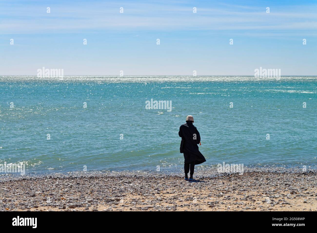 Young woman dressed in black with back to camera standing on shoreline looking out to an empty seascape at Birling Gap East Sussex England UK Stock Photo