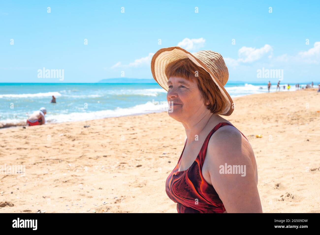 An adult elderly woman in a hat and a swimsuit on the sandy coast looks ...