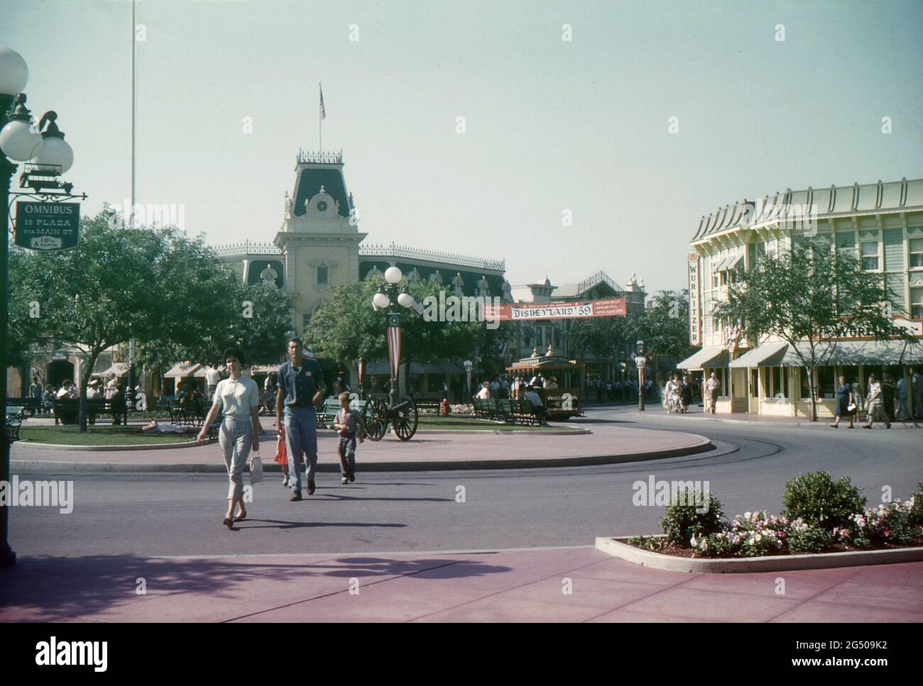 Disneyland, California, 1959. A view of Town Square and Main Street. A family are crossing the street heading towards Main Street Station, while a Disneyland horse-drawn street car is parked in the background. A sign for the Disneyland Omnibus is mounted on a lamp post. The Wurlitzer Music Hall can be seen on the corner of Main Street and the Town Square. A “Disneyland ‘59” banner is displayed across Main Street, advertising the resort’s new additions for that year. An antique cannon is on display in the square while visitors relax on benches and pass by. Stock Photo