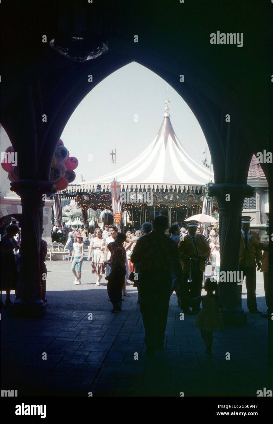 Disneyland, California, 1959. A busy scene showing a view of the King Arthur Carrousel from within Sleeping Beauty’s Castle, Fantasyland. Visitors are passing by and a balloon seller and Disneyland musicians are in the foreground. Stock Photo