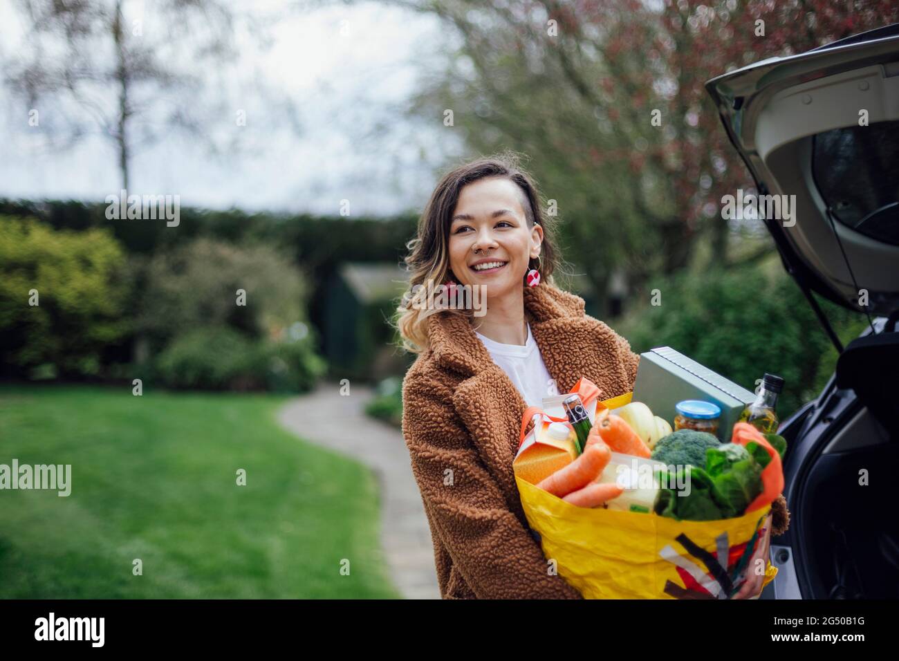 A young woman smiling, looking away from the camera and holding a reusable carrier bag filled with groceries. She is unloading the car. Stock Photo