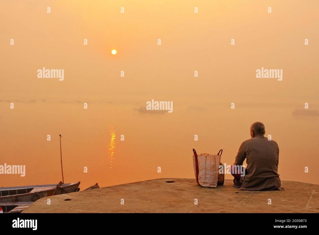 Old man meditates at Varanasi Ganges river bank at sunrise with view of wooden boats and ancient architecture. Stock Photo