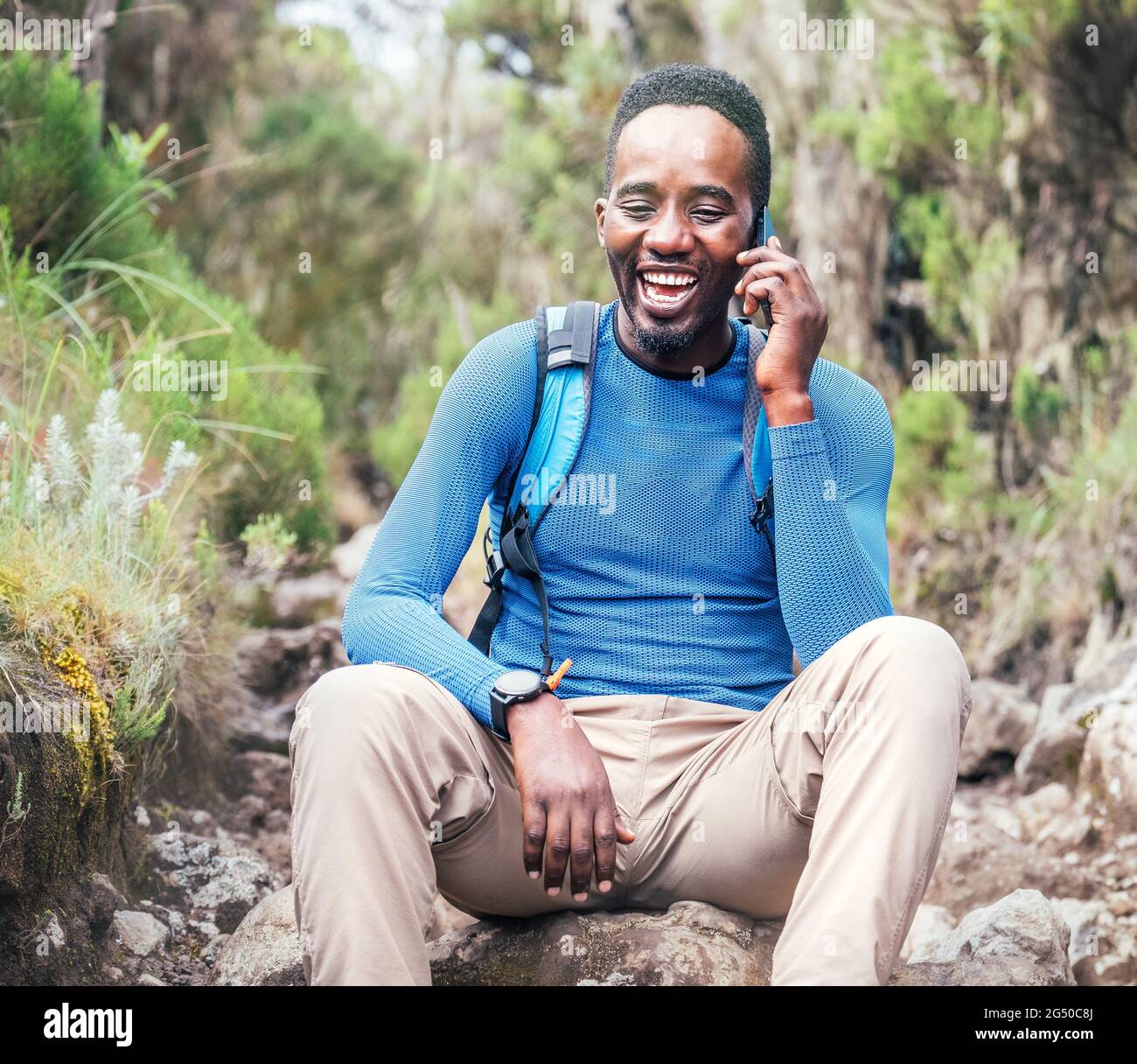 Young African ethnicity man talking via cellphone with somebody and cheerfully laughing as he having a hiking walk in the tropical forest. Happy peopl Stock Photo