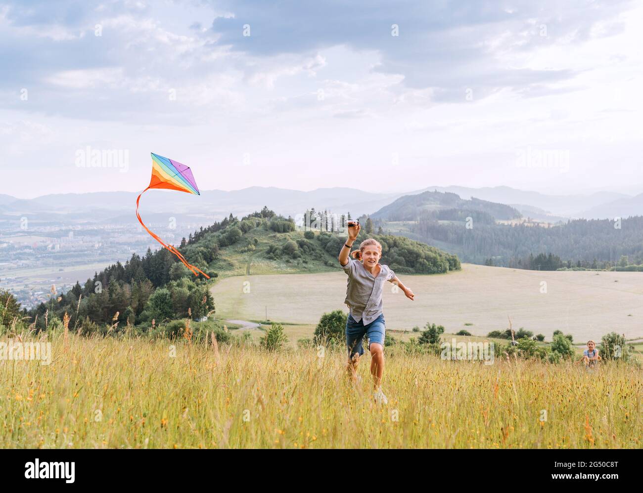 Smiling teenager boy with flying colorful kite on the high grass meadow in the mountain fields. Happy childhood moments or outdoor time spending conce Stock Photo