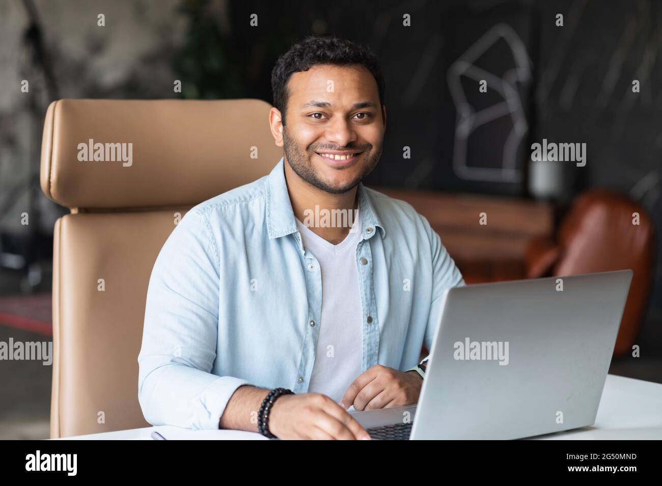 Optimistic confident dark-haired indian handsome guy wearing casual shirt using laptop for work in the office, mixed-race eastern freelancer man sits at the desk and looks at camera, smiles friendly Stock Photo