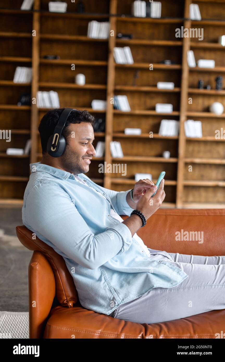 Attractive young indian hipster man wearing headphones relaxing and lying down on the comfy sofa holds smartphone listening music chatting in social media on mobile phone watching movies Stock Photo A...