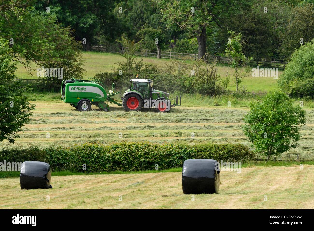 Hay or silage making (farmer in farm tractor at work in rural field pulling baler, collecting dry grass & round bales wrapped - Yorkshire England, UK. Stock Photo