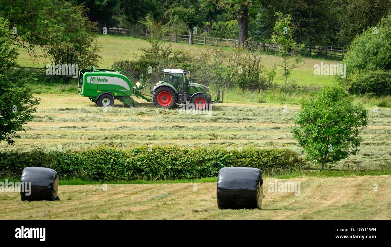 Hay or silage making (farmer in farm tractor at work in rural field pulling baler, collecting dry grass & round bales wrapped - Yorkshire England, UK. Stock Photo