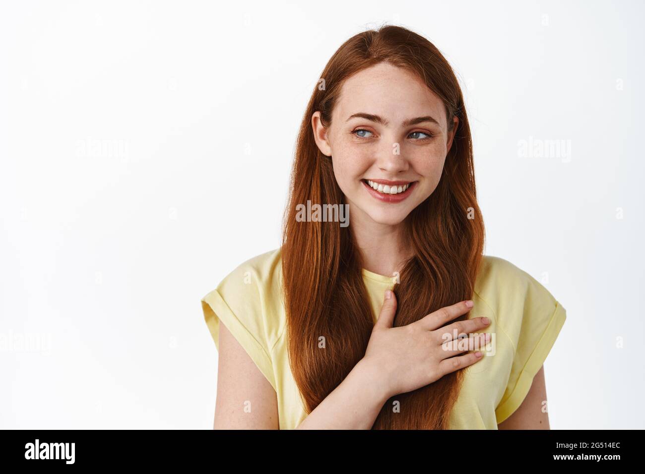 Tender and cute redhead female model holding hand on heart, looking aside at logo with heartwarming emotion, feel romantic, daydreaming, white Stock Photo