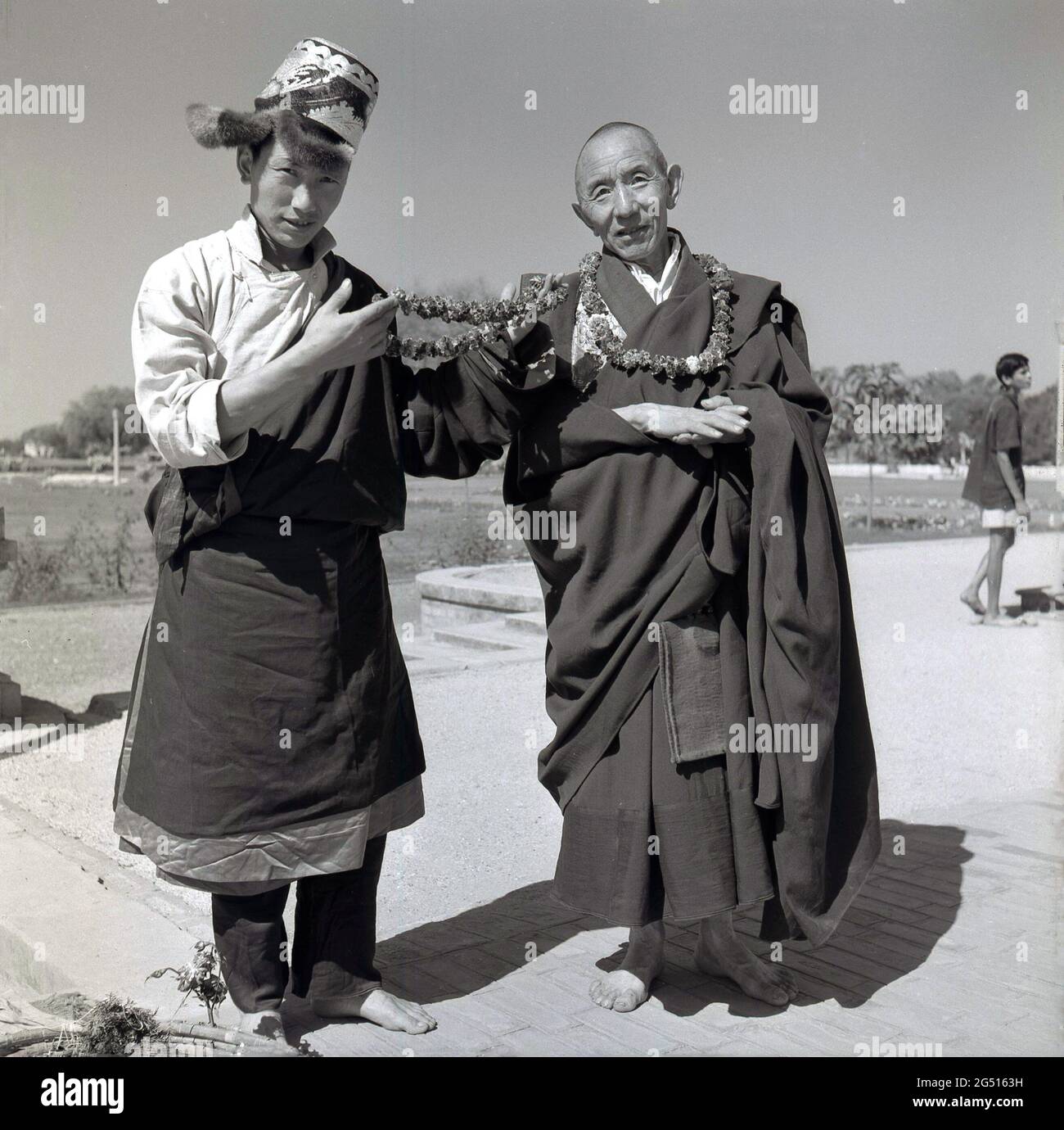 1950s, historical photo by J Allan Cash of a youth in robes and hat, standing with a monk outside the Buddhist temple, mulagandhakuti vihara, Sarnath, Varanasi, india. The young man is holding flower garlands or braids, which are worn around the neck and which he is offering to visitors. The temple was built in 1931 - on the site where the Buddha Gautama mediated during his first rainy season - by Angarika Dharmapala and is maintained and run by the Mahabodhi Society of which he founded. Stock Photo