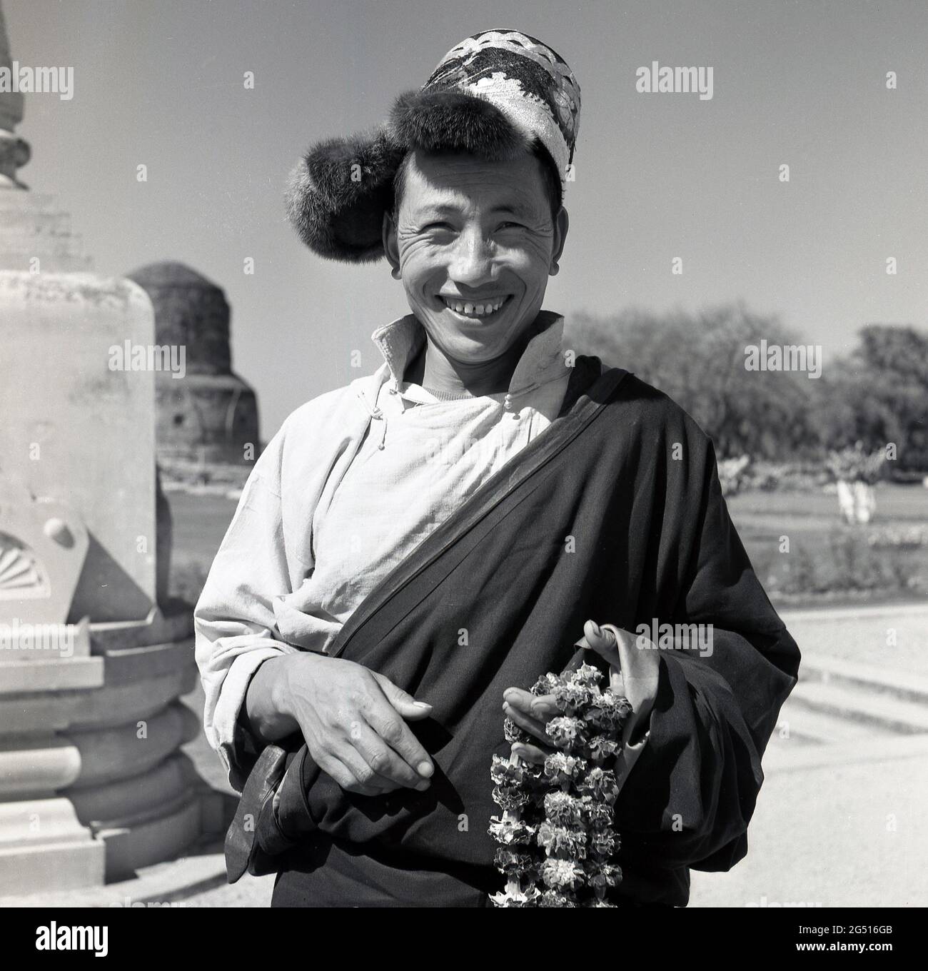 1950s, historical photo by J Allan Cash of a smiling, happy youth standing outside the Buddhist temple, mulagandhakuti vihara, Sarnath,Varanasi, india, holding flower garlands or braids, which are worn around the neck and which he is offering or selling to visitors. The temple was built in 1931 - on the site where the Buddha Gautama mediated during his first rainy season - by Angarika Dharmapala and is maintained and run by the Mahabodhi Society of which he founded. Stock Photo
