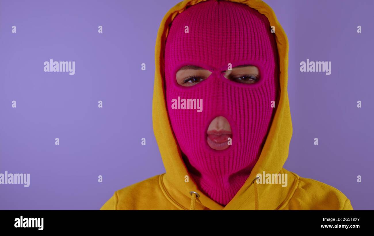 Close up of unrecognizable woman in pink balaclava shows her tongue on purple background. Unknown female in mask sticks out tongue and hides it, looki Stock Photo