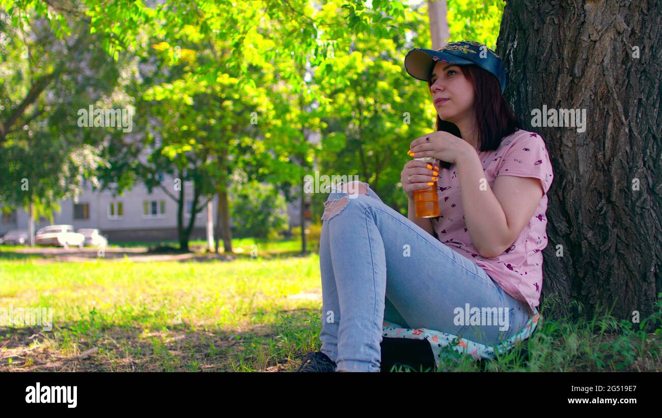 Young woman rests and drinks apple juice, sitting near tree on street