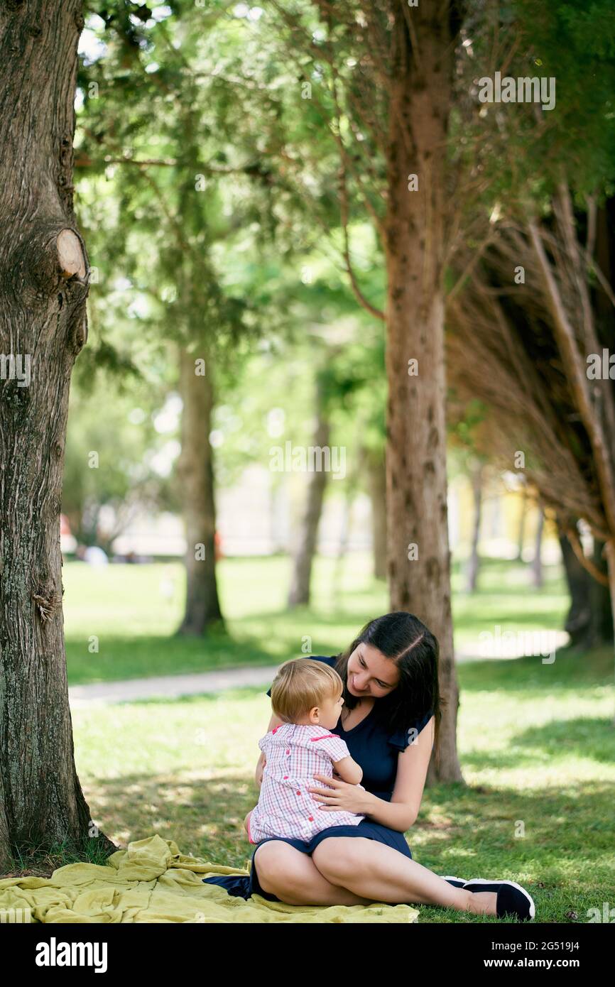 Mom holds on her lap a little girl sitting on a blanket on the lawn