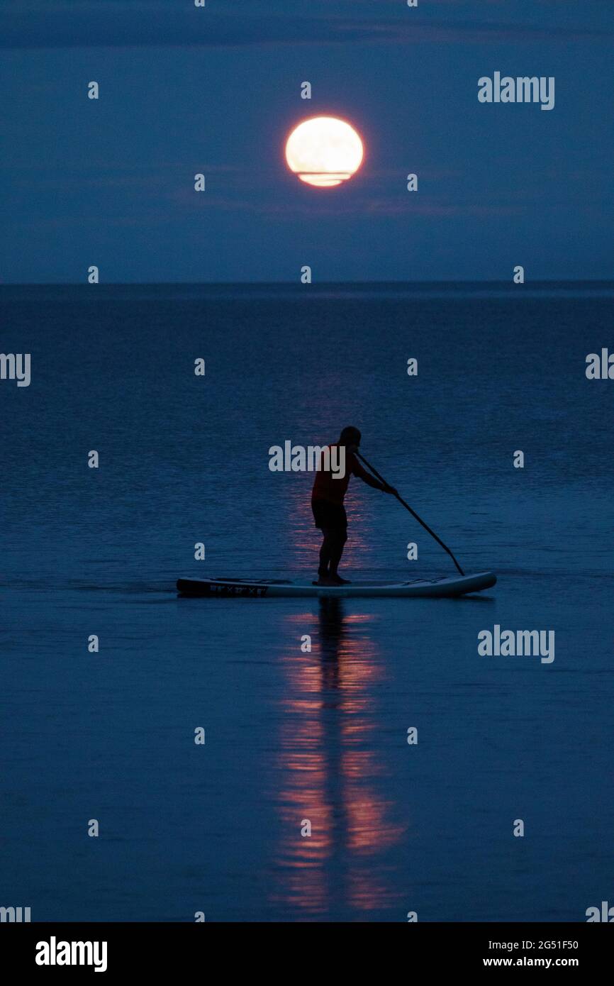 Cawsand Bay, Cornwall, UK. UK Weather: 24th June 2021. The Strawberry Full Super Moon rising on the horizon at Cawsand Bay with clearing sky to view the spectacle as this paddle boarder discovered © DGDImages/AlamyNews Stock Photo