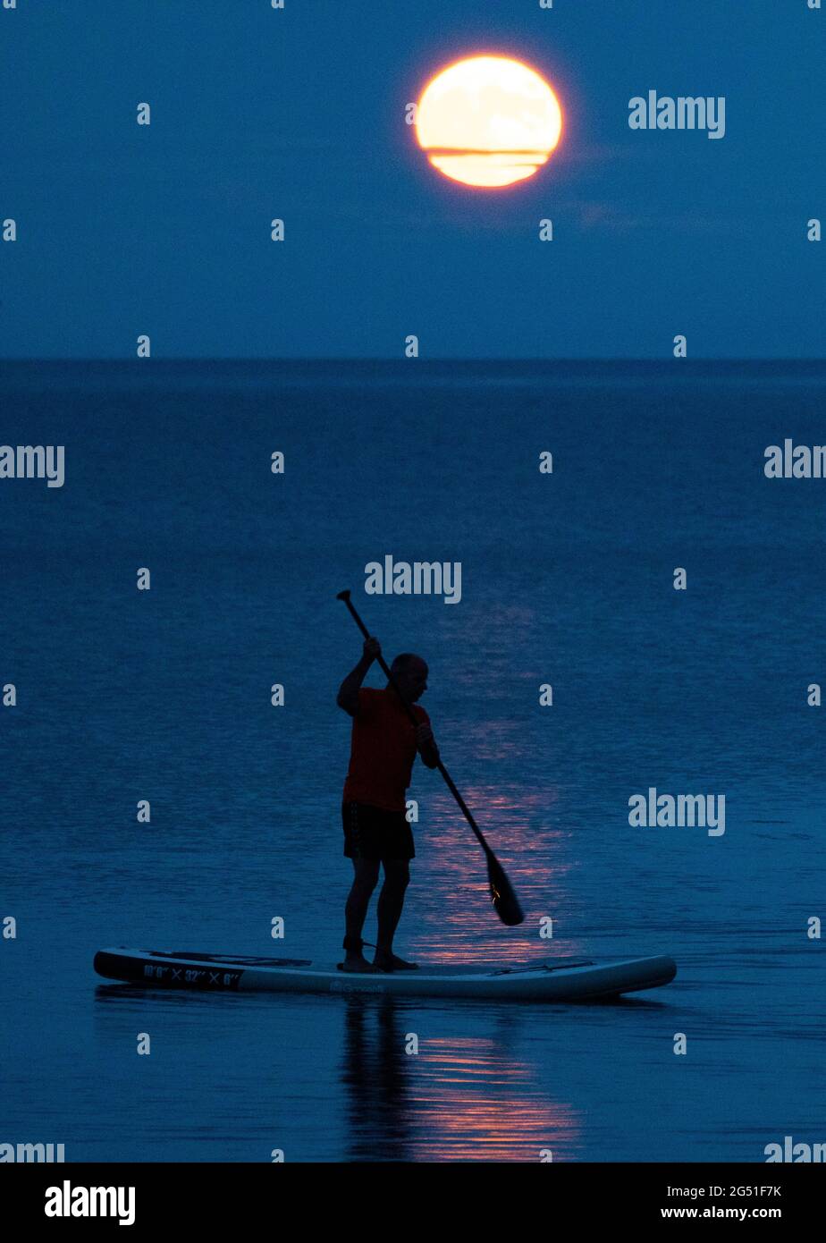 Cawsand Bay, Cornwall, UK. UK Weather: 24th June 2021. The Strawberry Full Super Moon rising on the horizon at Cawsand Bay with clearing sky to view the spectacle as this paddle boarder discovered © DGDImages/AlamyNews Stock Photo