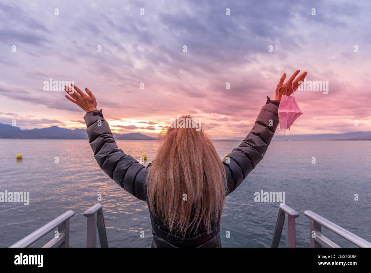 Unrecognized blonde woman in the coast raising her arms and holding her face mask. Horizontal photo Stock Photo