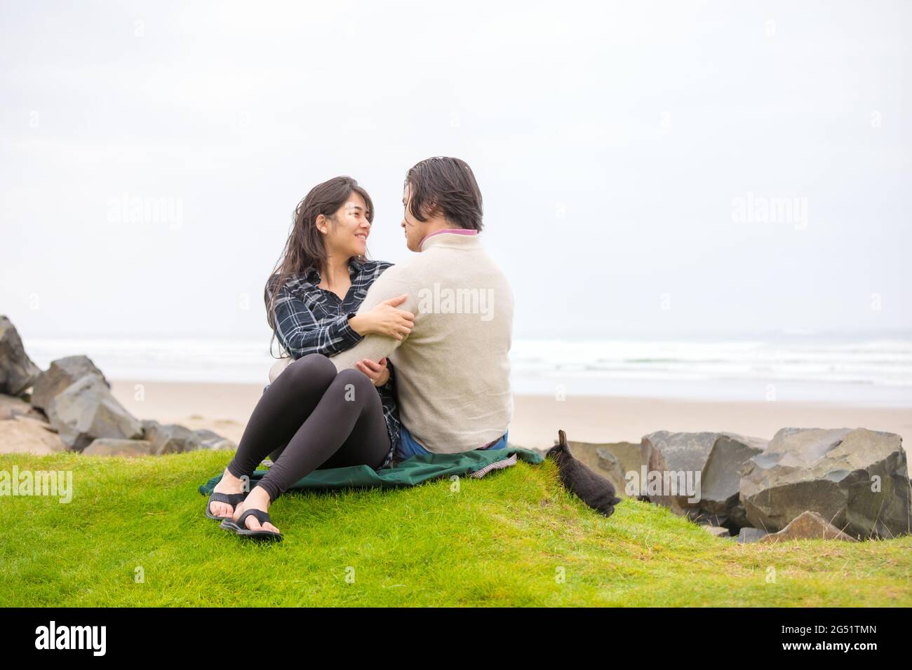 Young biracial couple sitting on grass hill over sandy beach next to ocean on foggy day Stock Photo