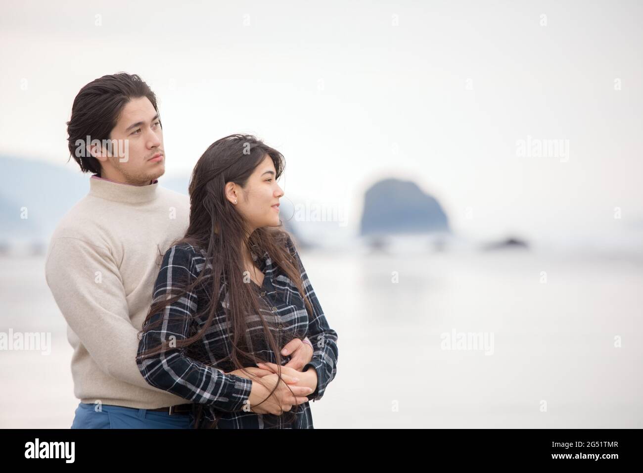 Biracial couple stabding on beach looking out towards ocean on foggy spring day Stock Photo