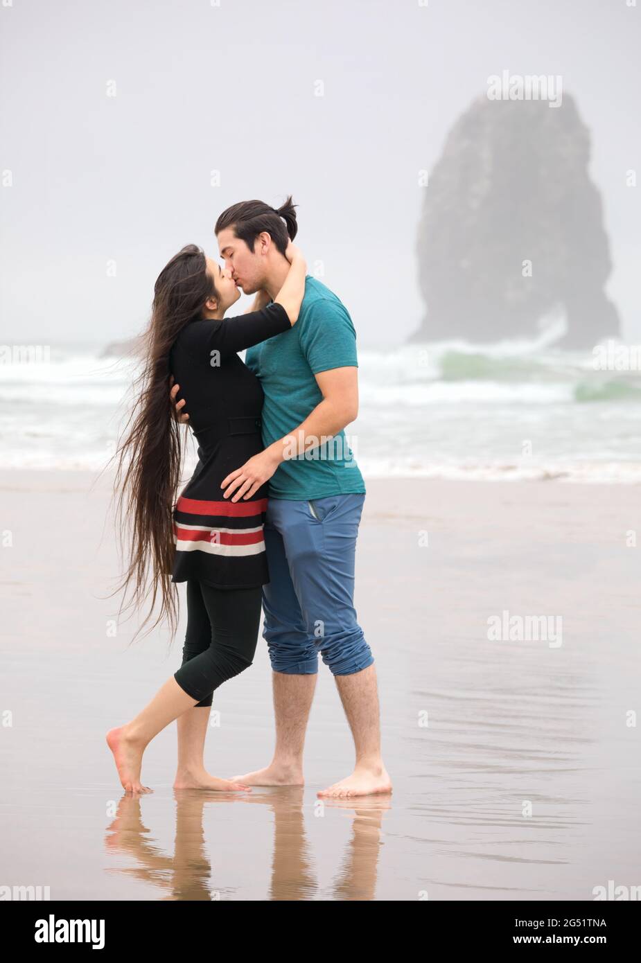 Biracial young couple kissing on beach by Pacific Ocean on foggy day, pants rolled up Stock Photo