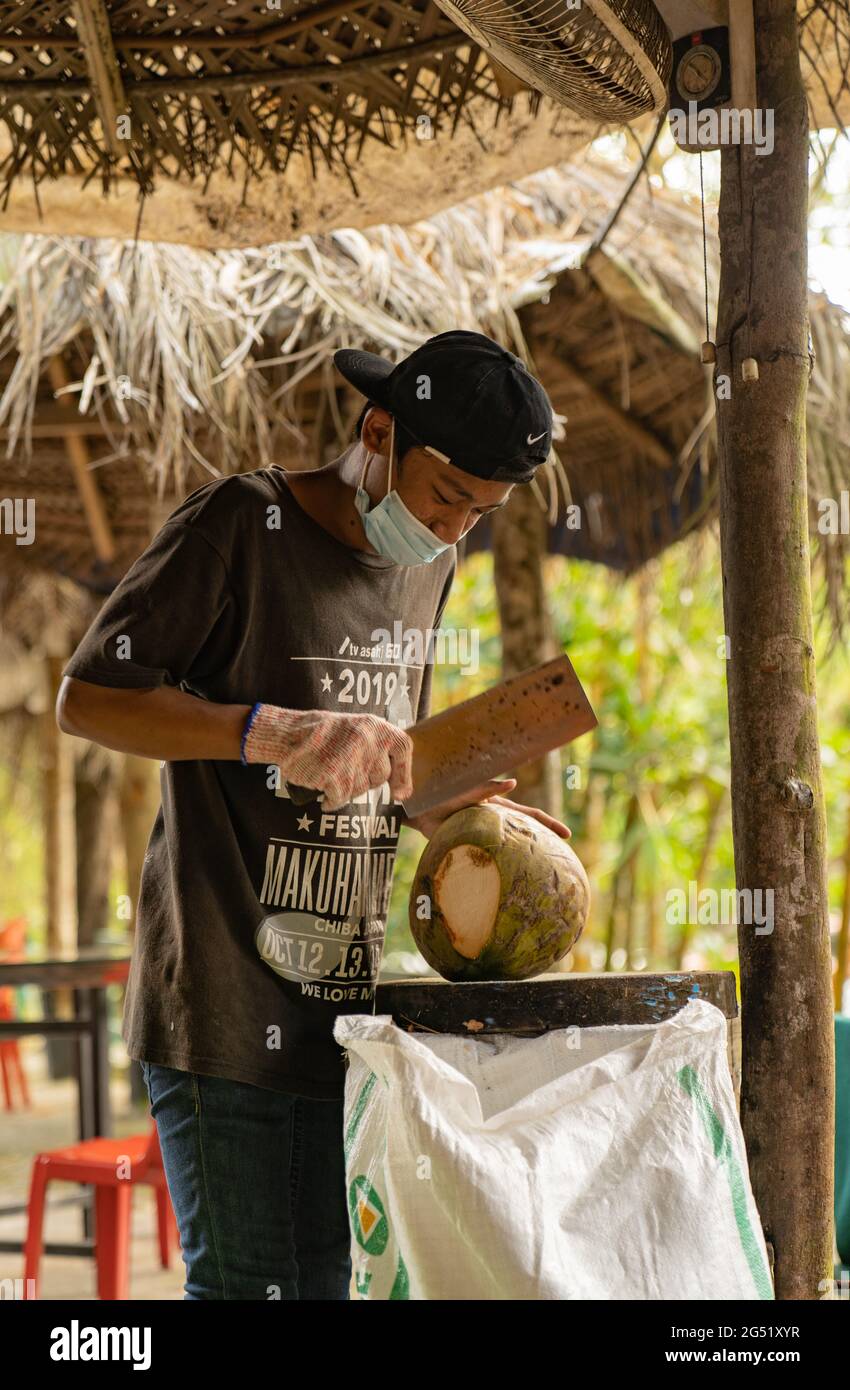 Asian boy wearing a mask opening coconuts with a big knife in a street market in Malaysia Stock Photo