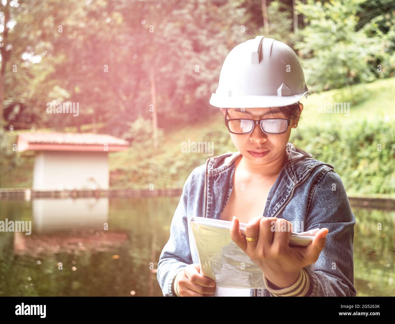 Female ecologist in safety hat working on site at the dam and stands reading the blueprints at the construction site. Stock Photo