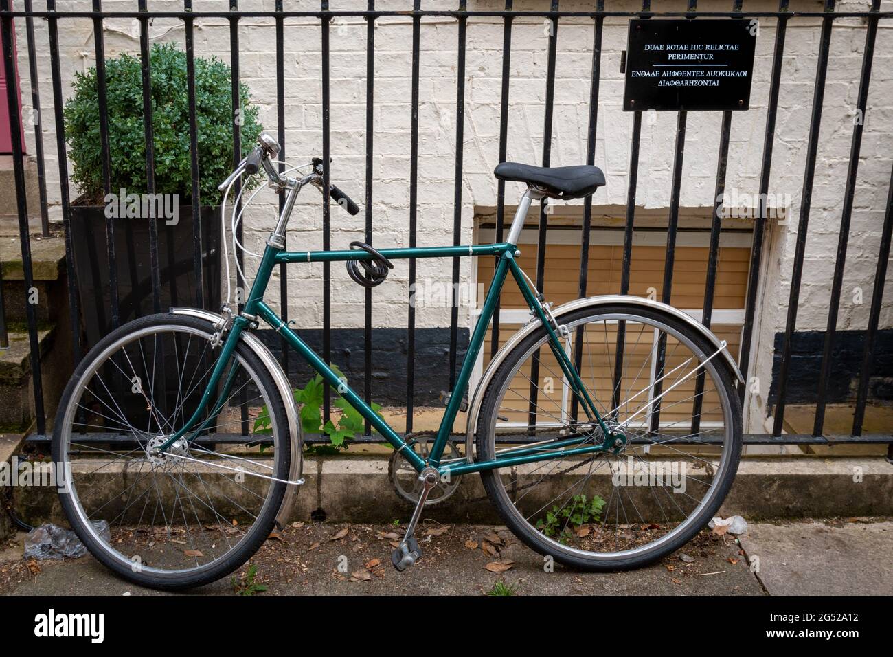 A bicycle is chained to a fence next to a sign asking people not to park their bicycles there in Latin and Greek. Cambridge, UK Stock Photo
