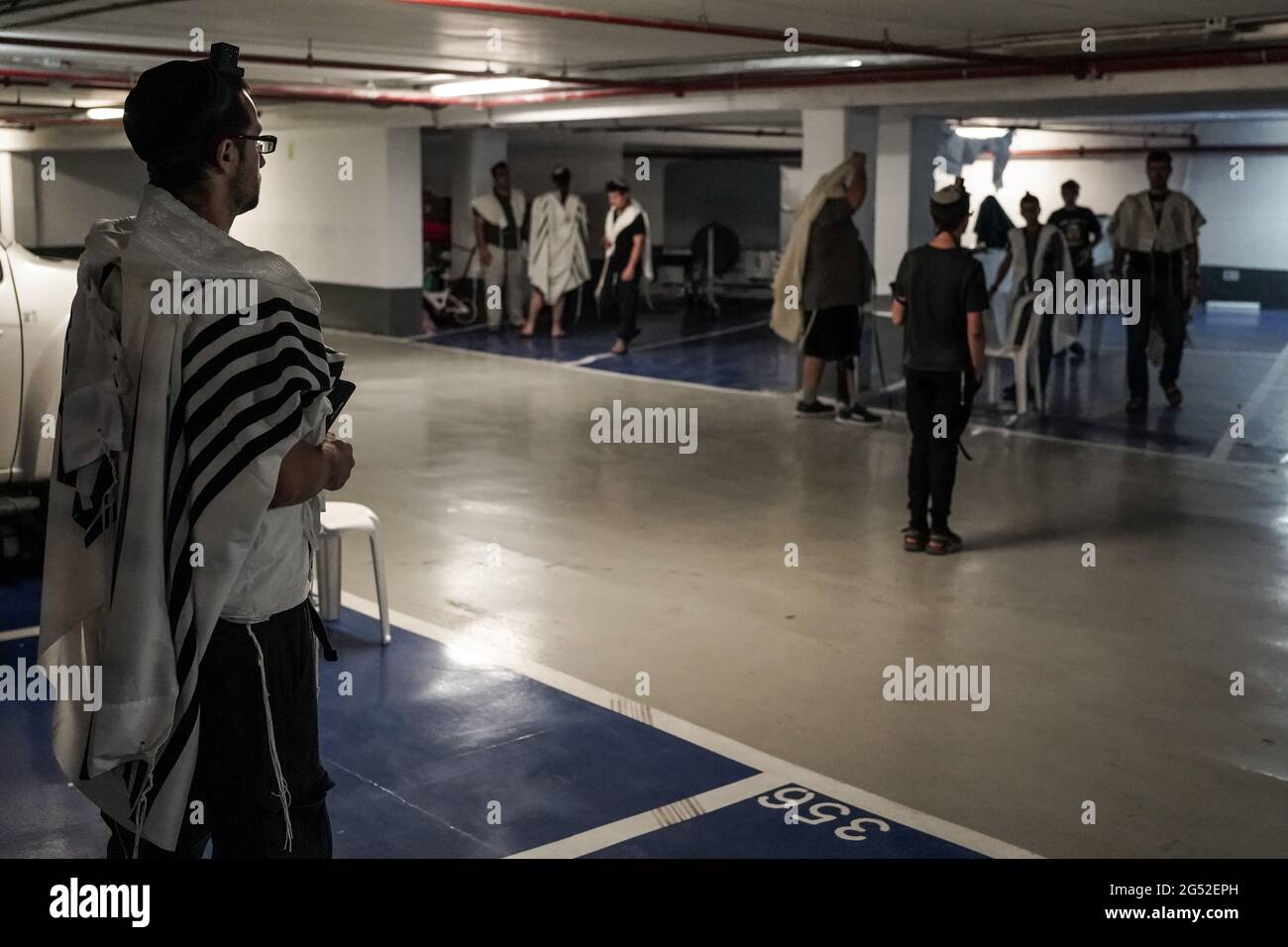 Jerusalem, Israel. 25th June, 2021. Jewish men, covered in prayer shawls, conduct their morning prayers in an underground parking lot in an act of self imposed social distancing, not mandatory at the moment. 138 new cases of COVID-19 were detected yesterday, over fifty percent among youth under the age of 15, the majority of type Delta Variant. PM Bennett announced the government is treating the increase in coronavirus cases as a new outbreak and taking measures to counter the disease. Face masks are once again mandatory indoors as of 12 noon today. Credit: Nir Alon/Alamy Live News Stock Photo