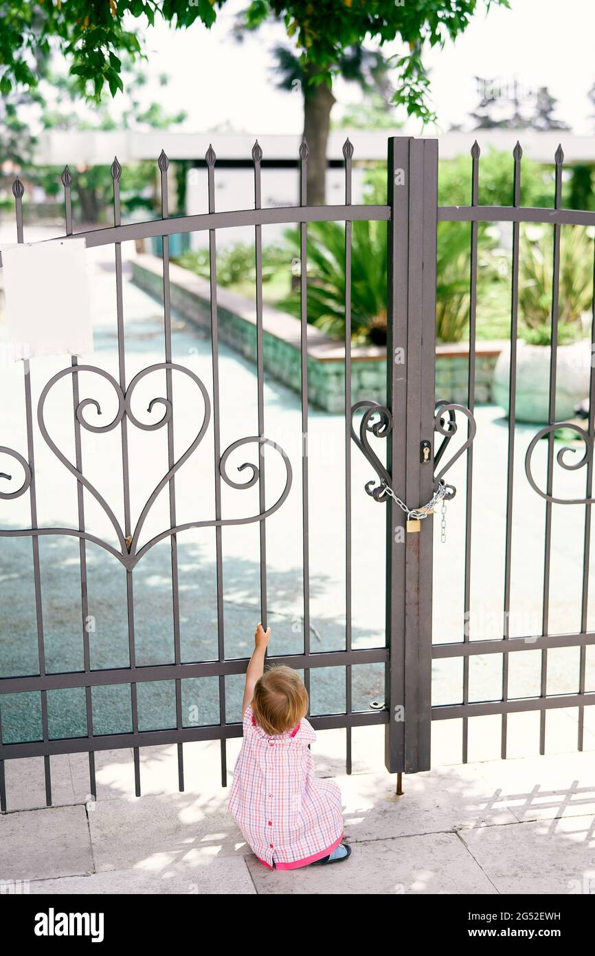 Little girl sits in front of a beautiful forged gate to the green park Stock Photo