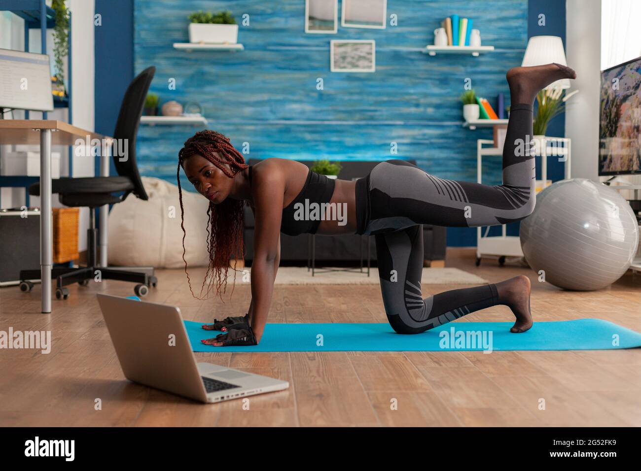 Black woman doing straight- leg donky kick pushing left foot up keeping leg bent, watching tutorial video on laptop screen, wearing sportwear on yoga mat for muscle strenght andn healthy lifestyle. Stock Photo
