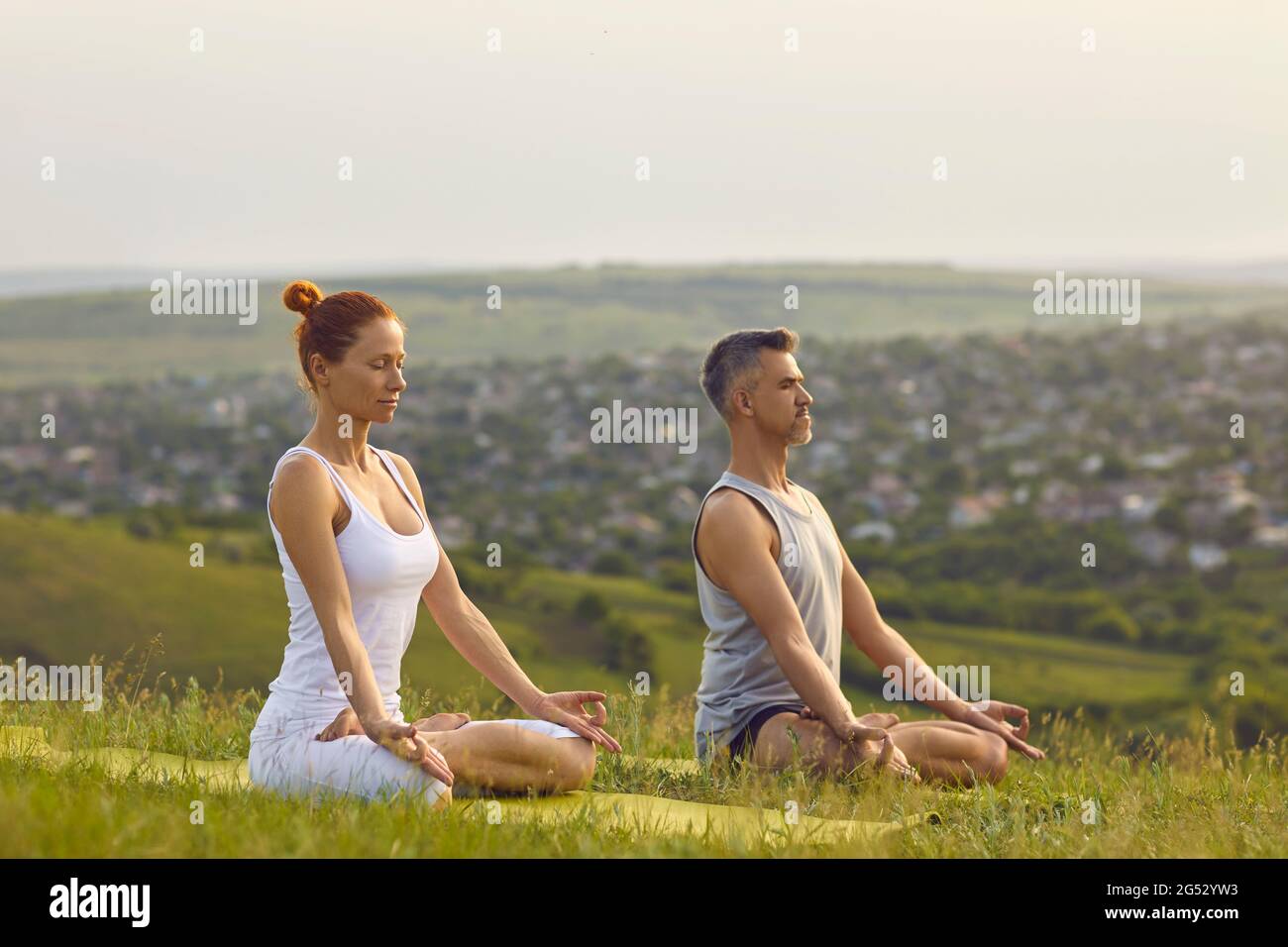 Middle aged man and woman breathing fresh air and meditating in Lotus pose on quiet green hill Stock Photo