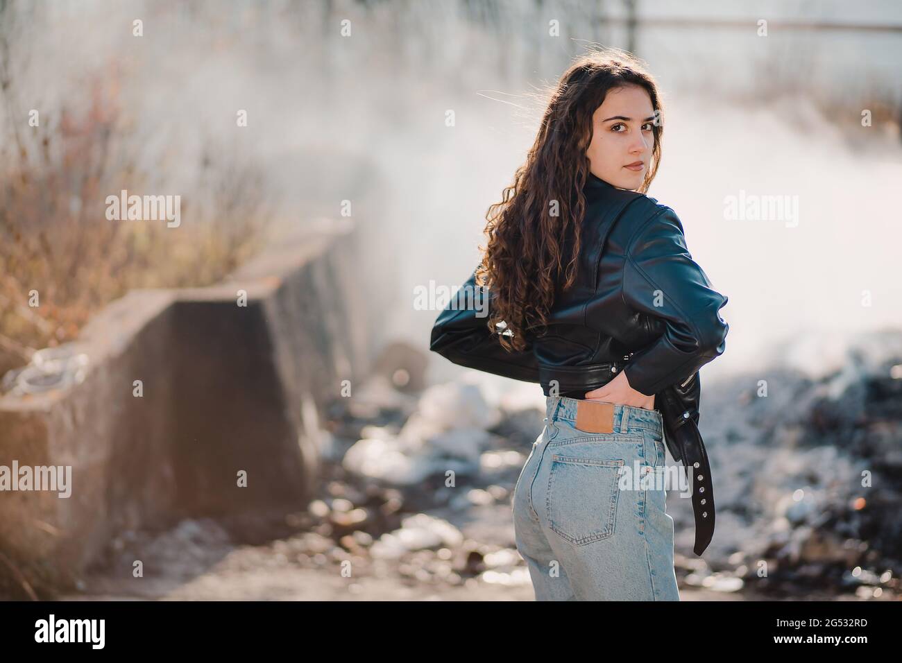 Outdoor portrait of a beautiful teen brunette girl in the black leather jacket with smoke in the background Stock Photo
