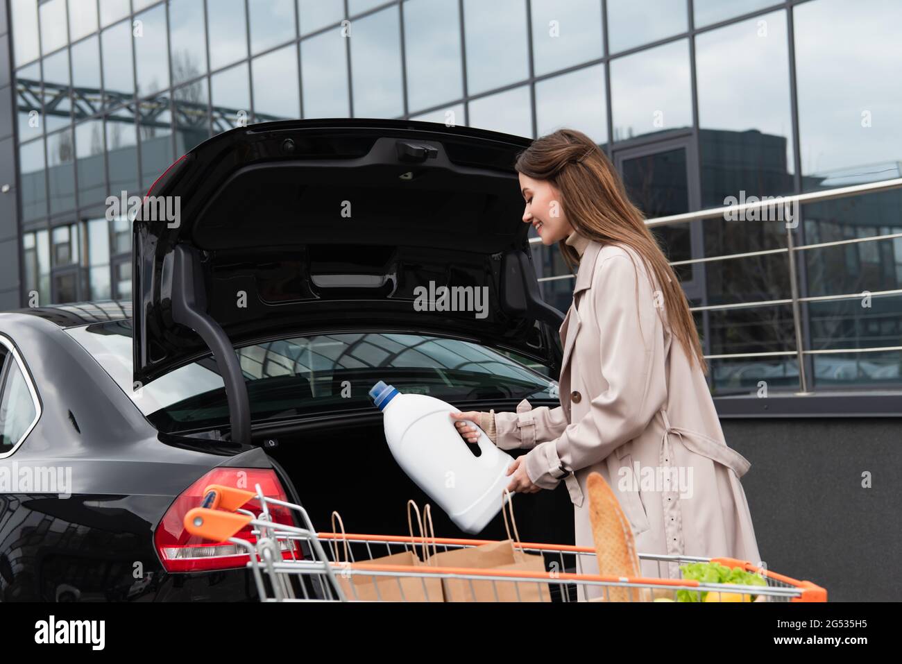 young woman putting bottle of liquid detergent into car trunk Stock