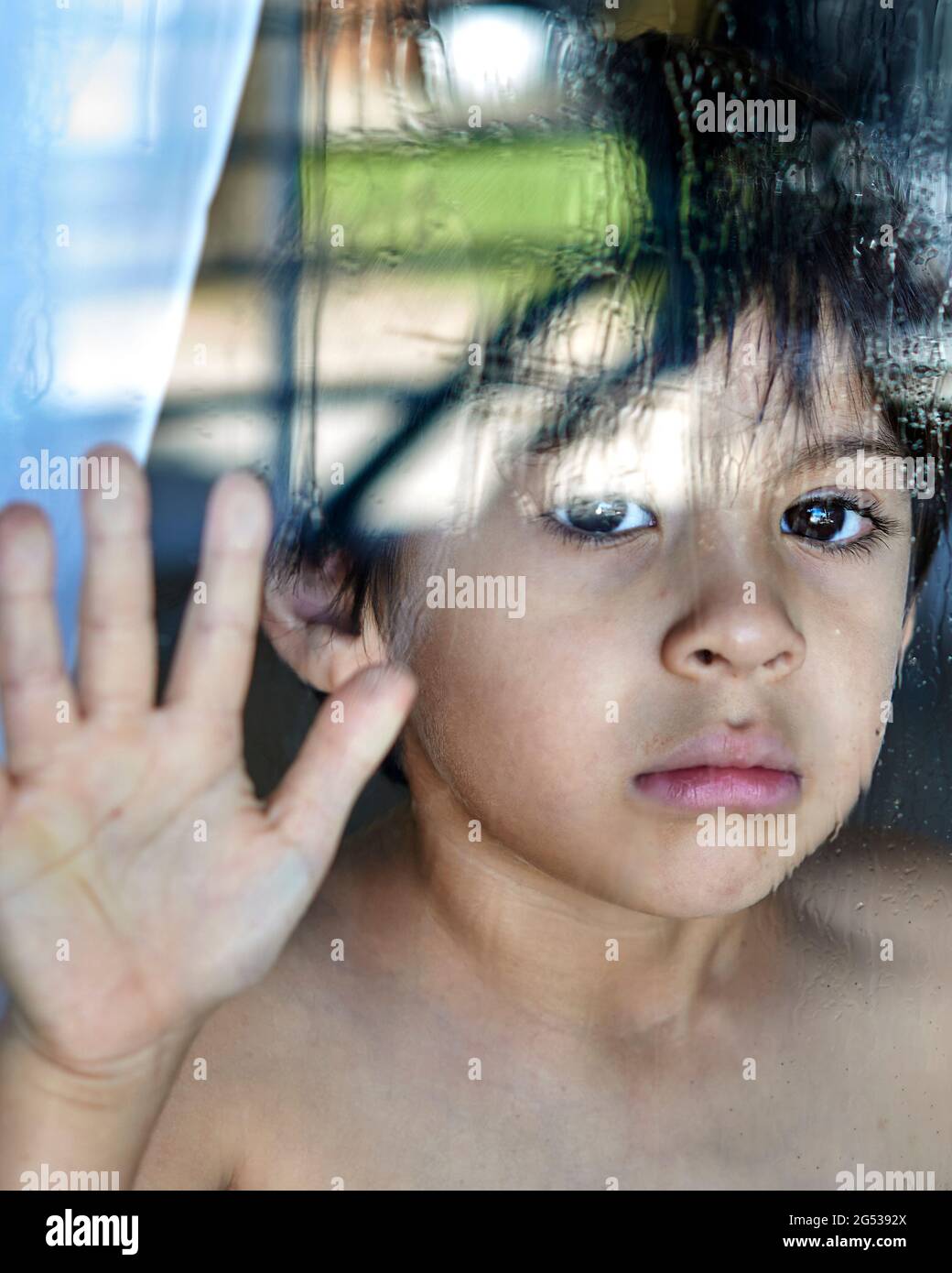 Title sad and anguished child looking behind the wet glass with hand resting on the window Stock Photo