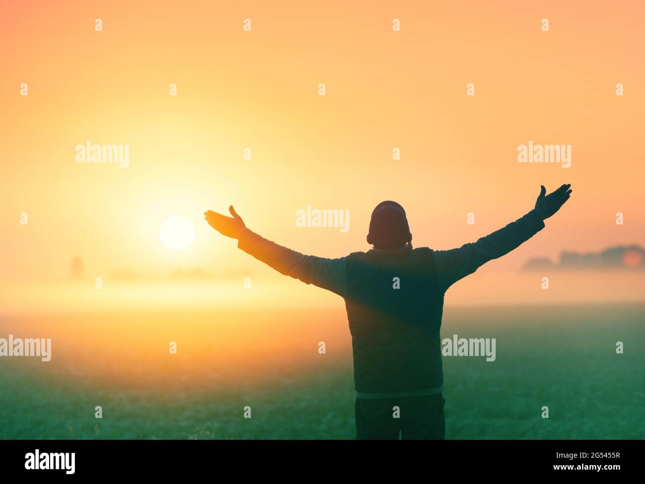 A man with raised arms stands in a field in the early morning and looks at the sunrise Stock Photo