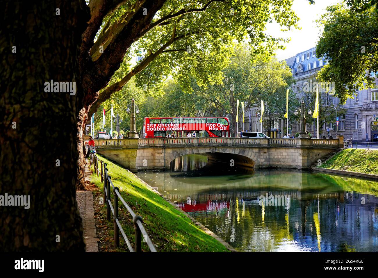 The beautiful city canal 'Stadtgraben' on Königsallee with its old trees. Red doubledecker tour bus waiting on a bridge crossing the canal. Stock Photo