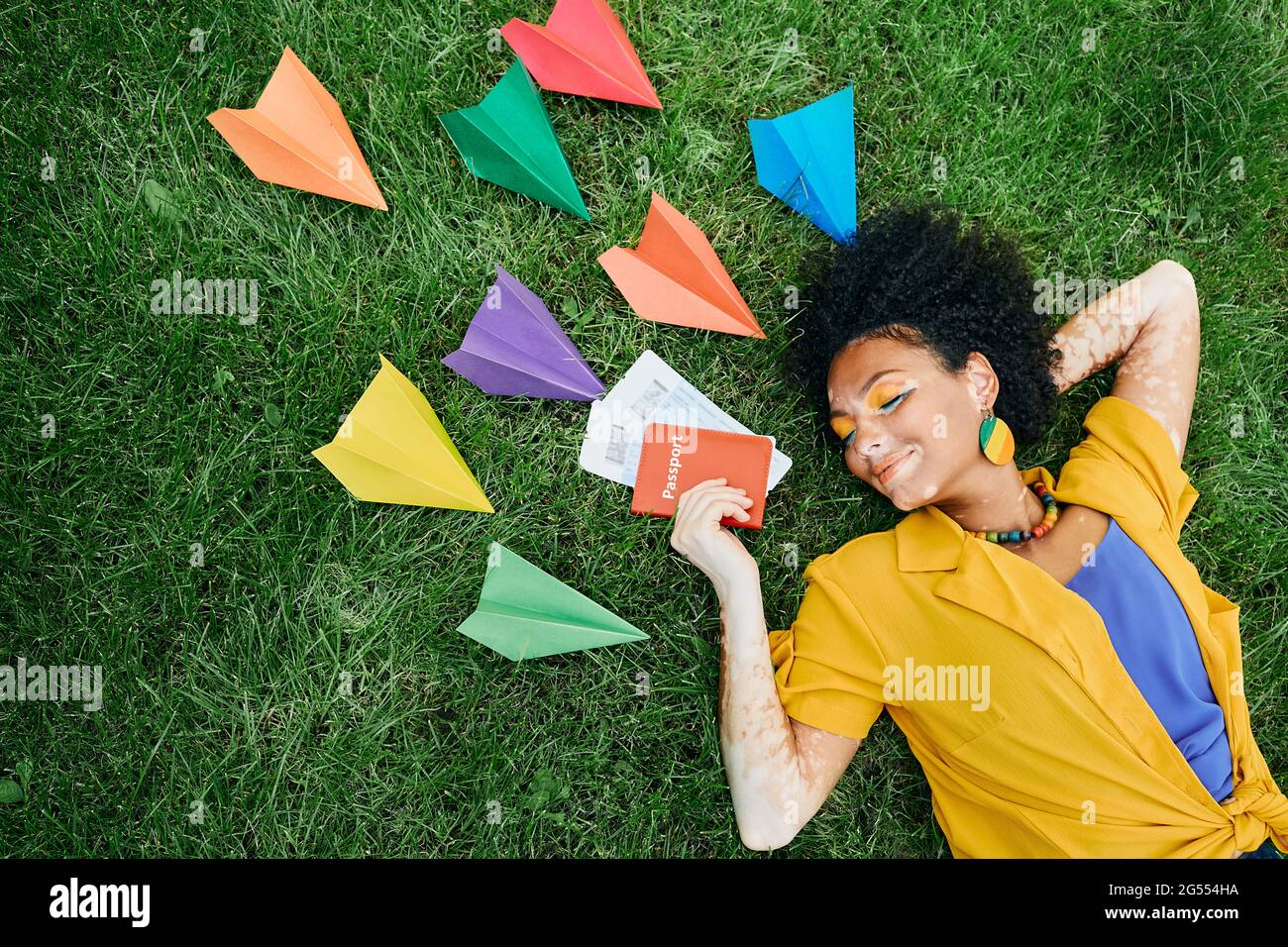 Traveling concept. Multi-ethnic woman with air tickets and international passport in hand dreams to vacations lying on green grass around colored pape Stock Photo