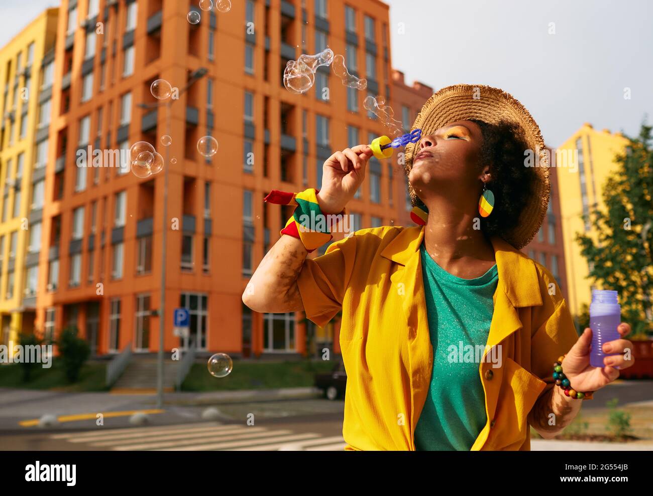 Stylish multi-ethnic woman blowing bubbles walking around modern city with colored buildings. Summer life in city Stock Photo