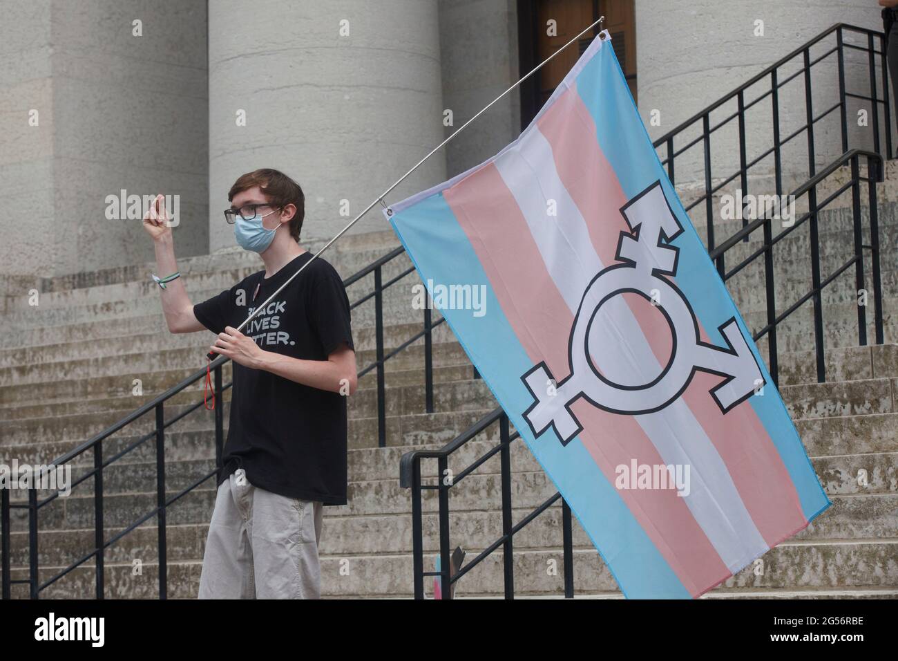 Ohio, USA. 25th June, 2021. A protester holds the trans flag and snaps in solidarity with other speakers, during the demonstration.Transgender rights advocates stood outside of the Ohio Statehouse at noon to oppose and bring attention to an amendment to a bill that would ban transgender women from participating in high school and college women sports. The original bill that this transgender ban was added to dealt with compensation for college students to profit off of their name, image and likeness. The addition of transgender ban to this bill was a surprise, because a transgender ban bill alr Stock Photo