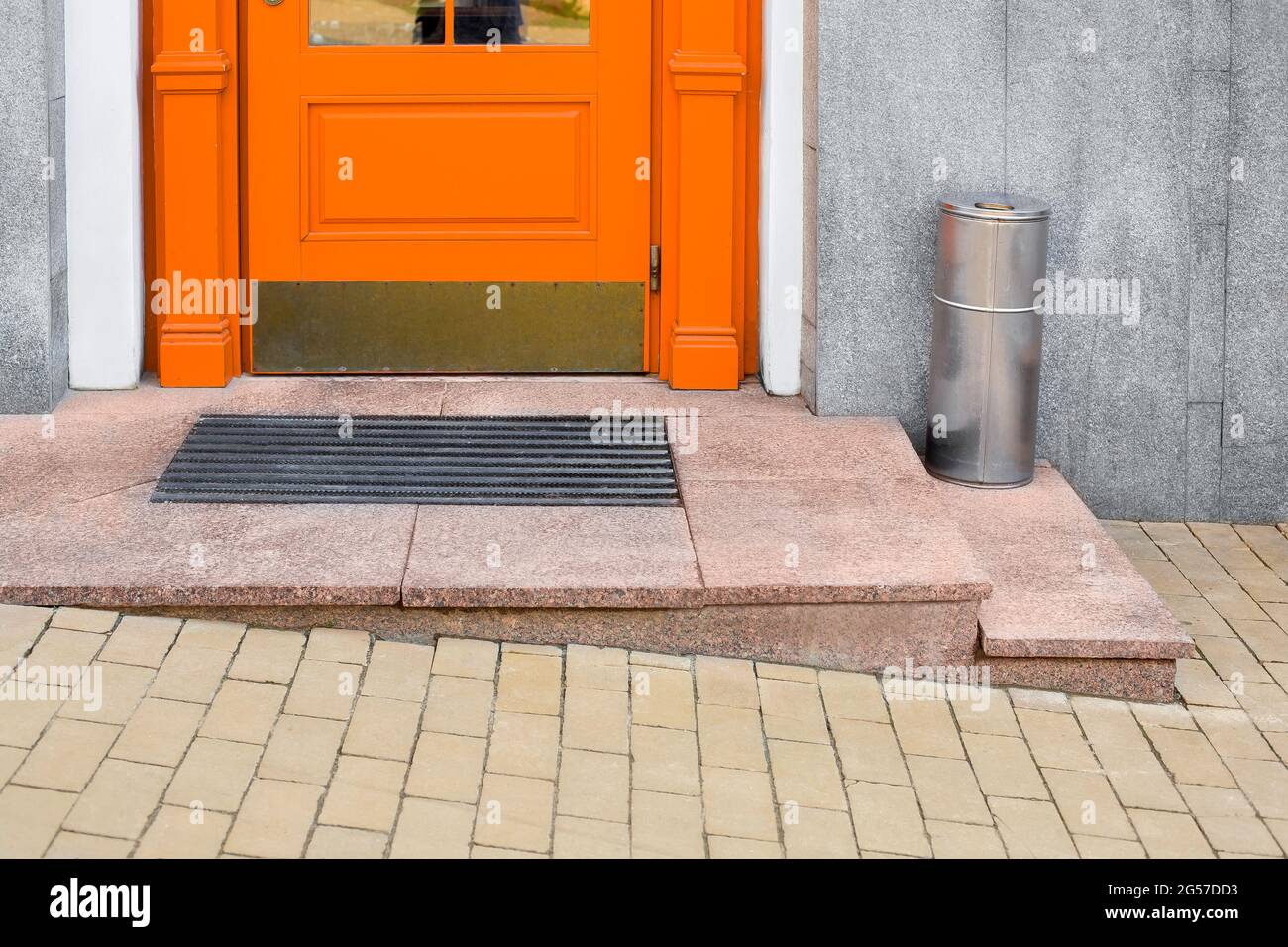 granite threshold with foot mat near orange wooden front door with iron trash can near building facade with gray cladding and yellow stone tile paveme Stock Photo