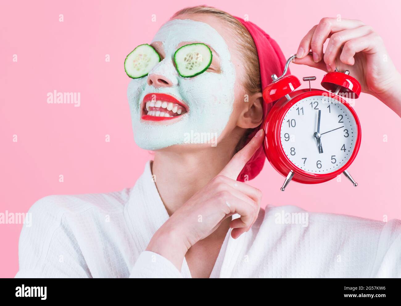 Smiling Woman with cosmetic facial mask and alarm clock in hand. Cosmetics procedure. Beauty spa and cosmetology. Stock Photo