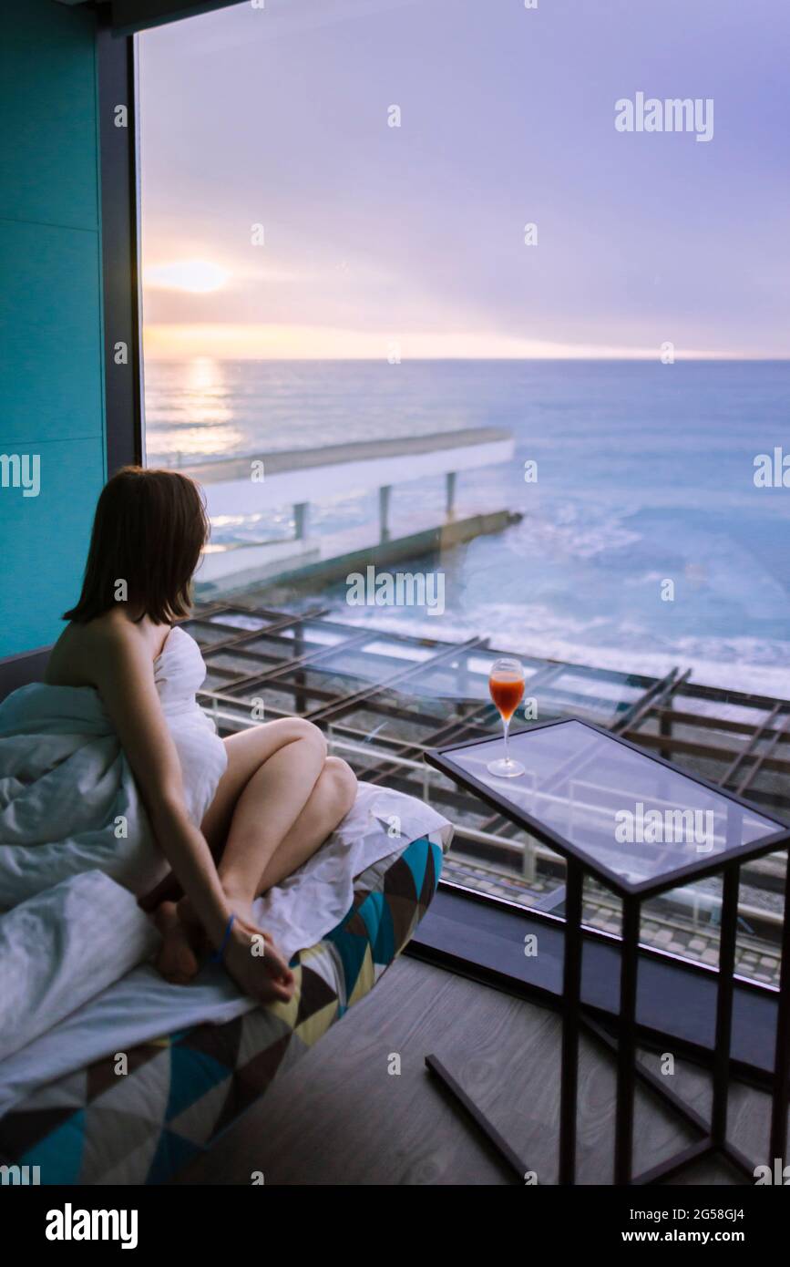 Woman sitting on bed with cocktail in hotel and observing sunrise over sea Stock Photo