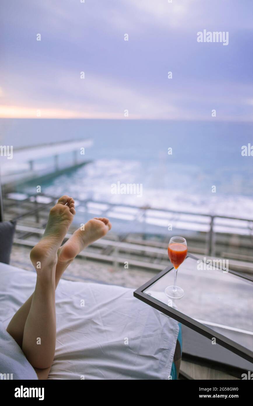 Woman's feet and cocktail in hotel room with view on sea at sunrise Stock Photo