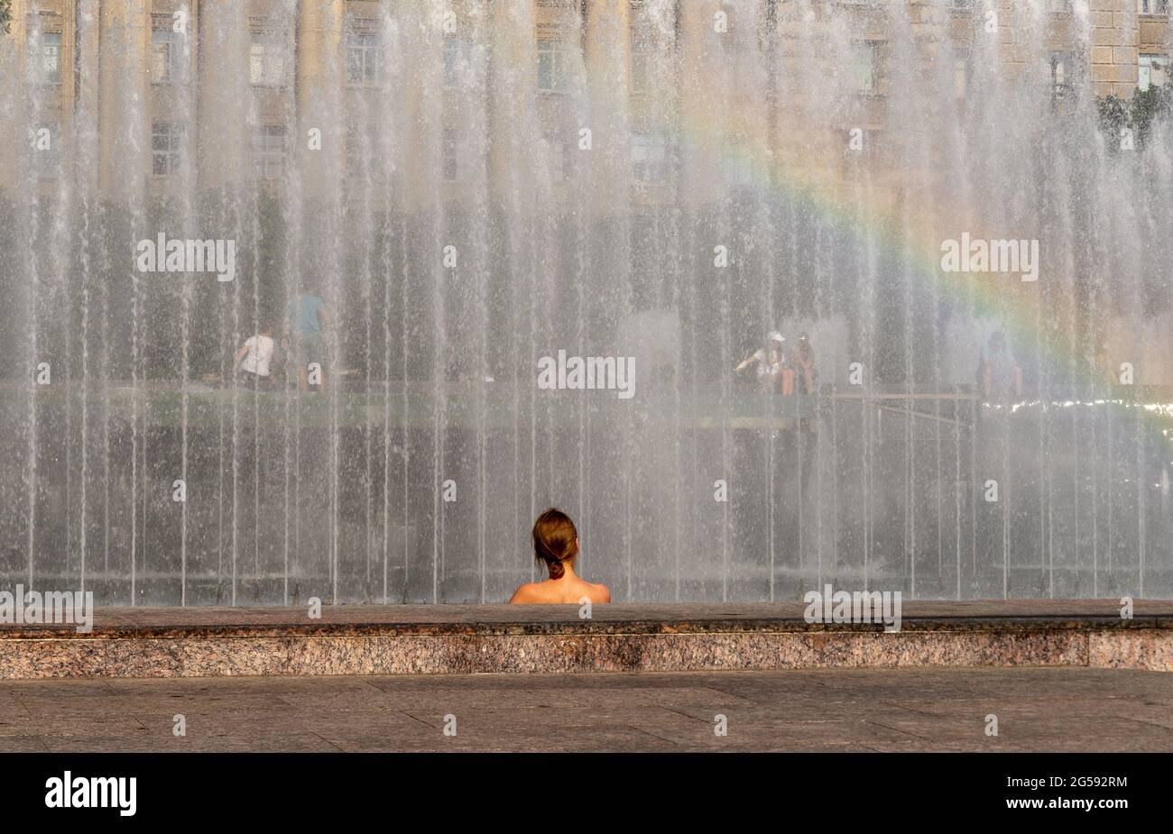 A woman's head and shoulders, back view, against the sprinkling fountain, with rainbow, on a hot summer day Stock Photo