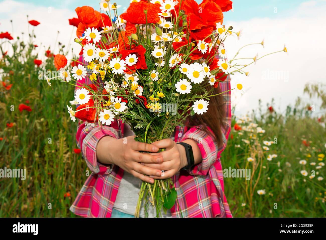 The girl stands against the background of wild flowers and holds a bouquet of poppies and daisies in her hands. A girl in the field was collecting a bouquet of wild flowers. Stock Photo