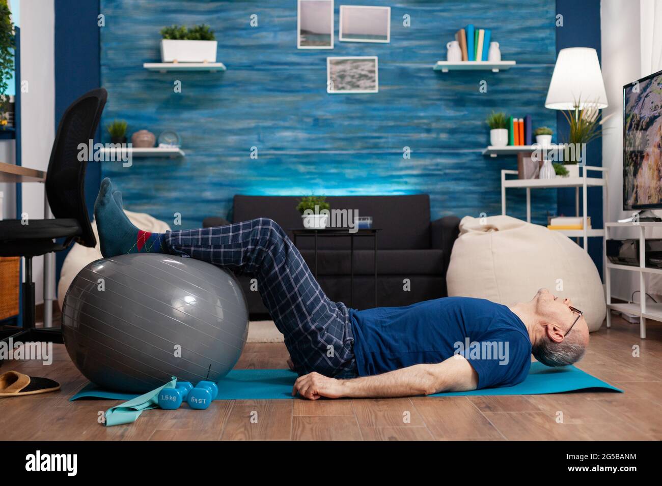 Retired senior sportman sitting on yoga mat practicing warming legs up exercises using swiss ball stretching abdominals muscles. Focused pensioner training body strength resistance in living room Stock Photo