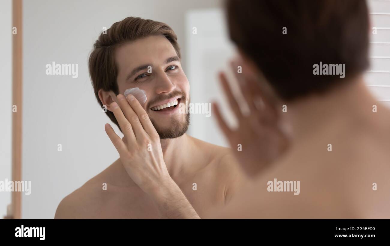 Happy handsome metrosexual guy applying sunscreen on face at mirror Stock Photo